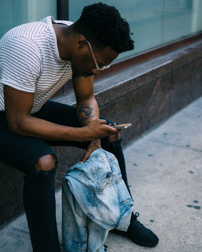 a young man sitting and leaning down to look at his mobile while outdoors