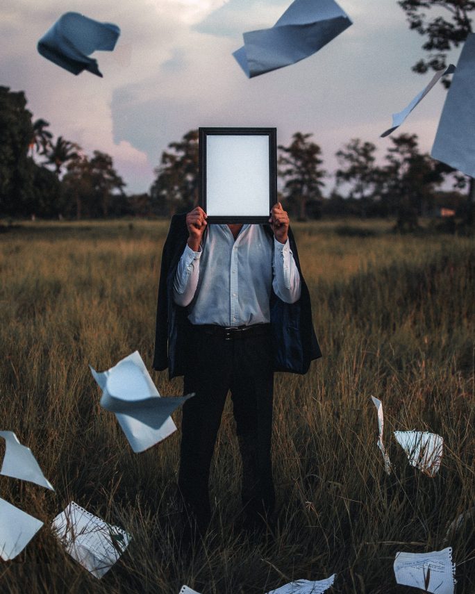 a person standing in a field holding a blank electronic tablet in front of their face with crumpled pages flying in the air around them