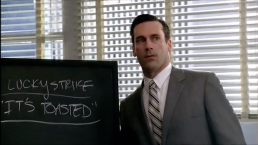 screenshot from the series Mad Men with Don Draper in front of a chalkboard that says Lucky Strike's It's Toasted