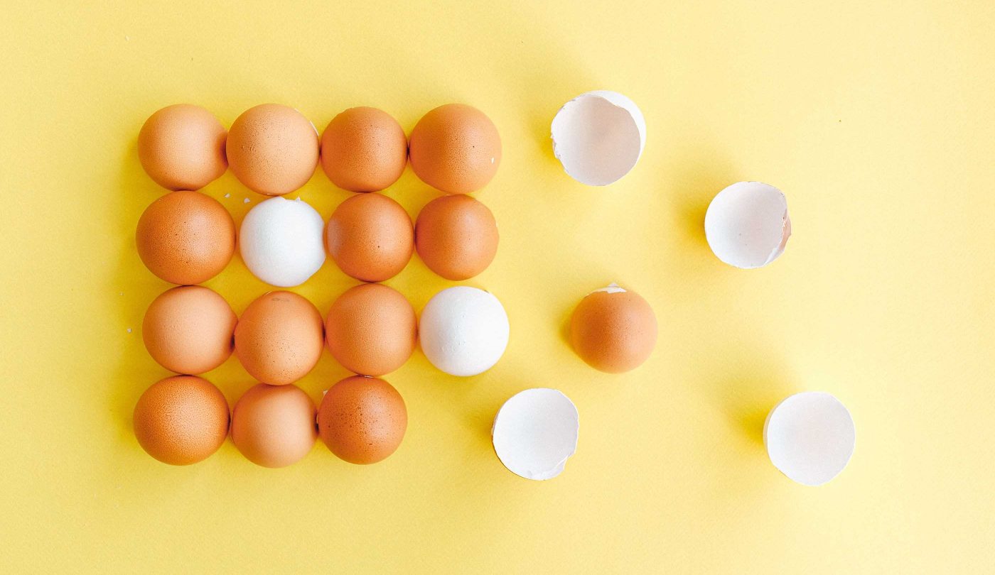 Brown eggs arranged in rows on a cheerful yellow background. In the second row, there is one white egg, providing a subtle contrast to the brown ones. On the right side of the rows, a couple of broken eggshells lay in halves.