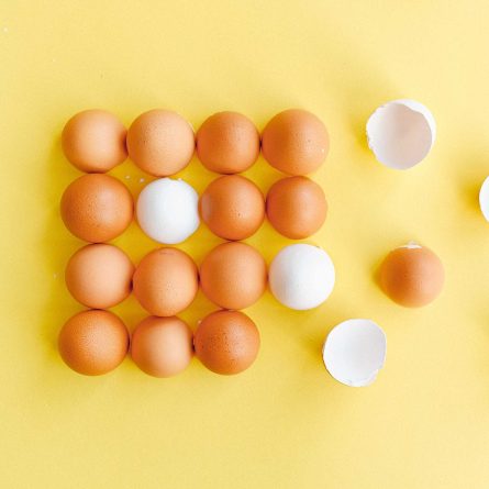 Brown eggs arranged in rows on a cheerful yellow background. In the second row, there is one white egg, providing a subtle contrast to the brown ones. On the right side of the rows, a couple of broken eggshells lay in halves.