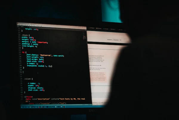 A computer screen in a dark room showing a coding interface is obscured by the shadowy figure operating it
