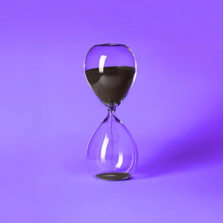 Clear glass hourglass with black sand partially emptied, set against a vibrant purple background.