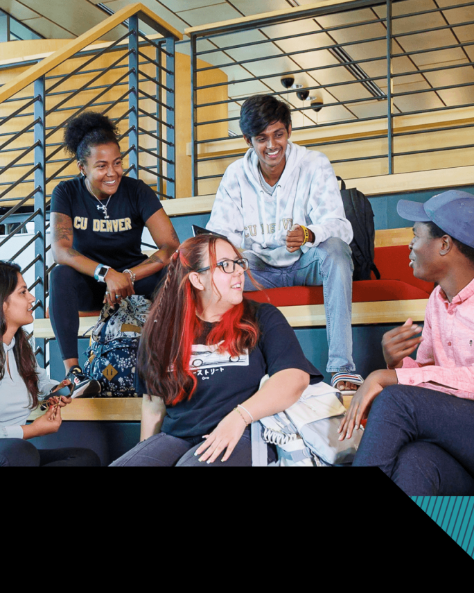 Diverse CU Denver students sit together on tiered seating inside a campus building, smiling and chatting in a relaxed, social atmosphere.
