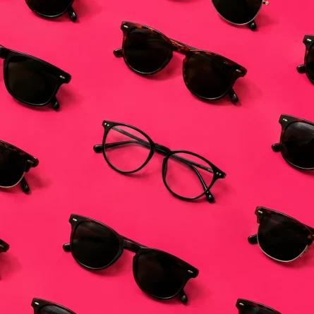 Assorted sunglasses surround a single pair of eyeglasses on a bright pink background, symbolizing visibility and standing out in AI-powered search.
