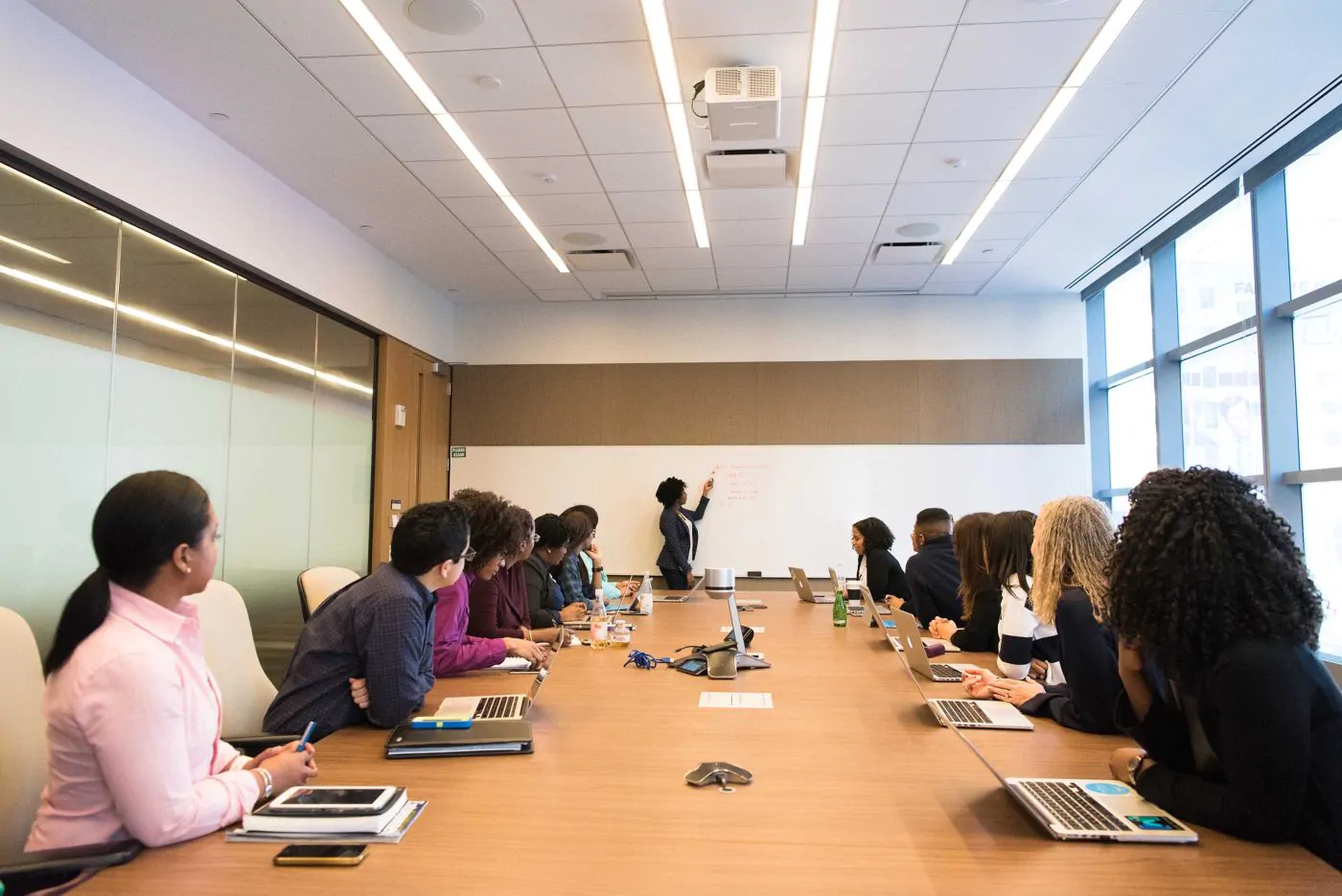 a board room of men and women looking at a woman writing on a white board
