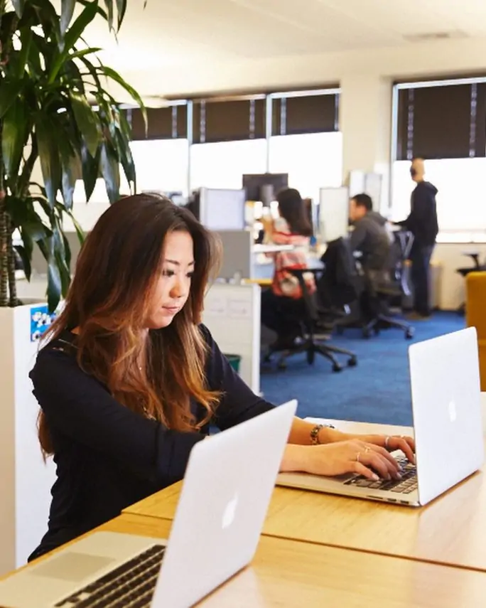 young woman typing on a laptop in a coworking space with other workers behind her in the distance