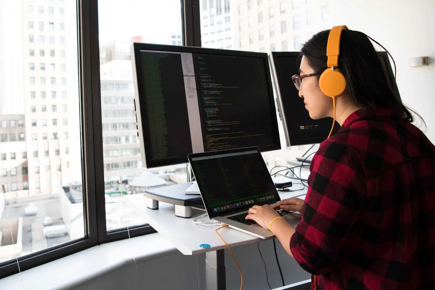 young woman working on code on three desktop monitors and one laptop