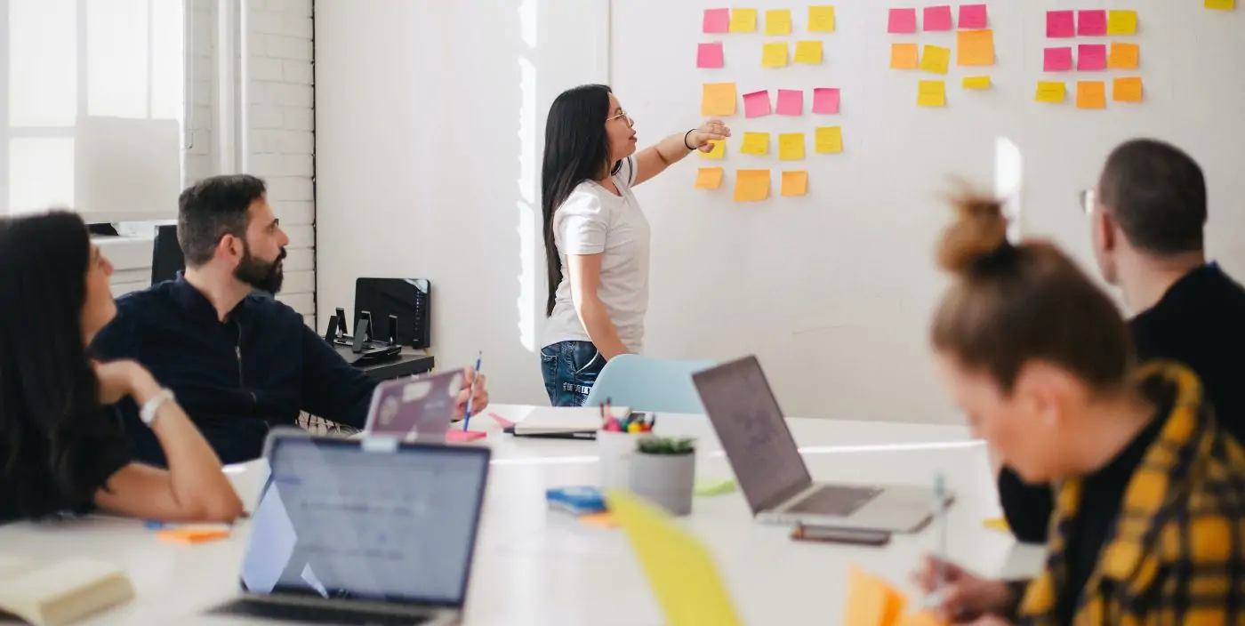 a young woman leading a white board session while other young women and men look on from a conference table with laptops