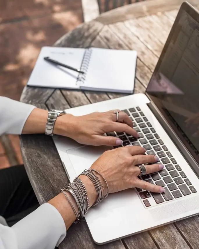 a woman's hands typing on a laptop keyboard and a notebook and pen next to her