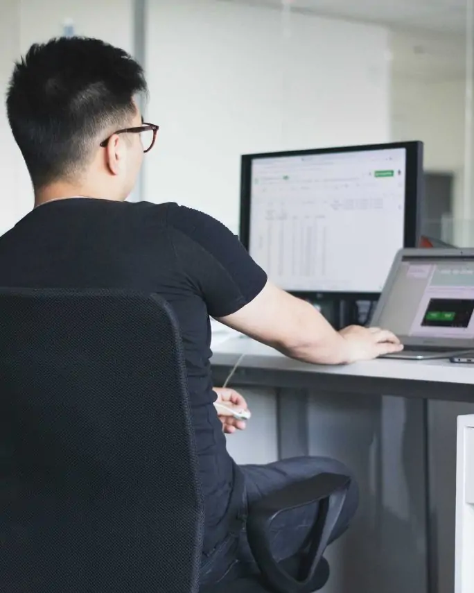 a young man working on two computer monitors, one of which has a spreadsheet full of data on it