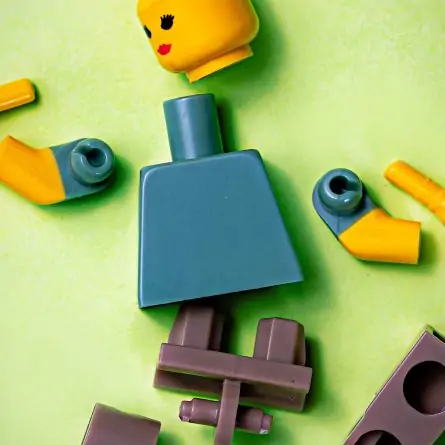 A broken LEGO person lying on a vibrant green background. The LEGO figure, composed of yellow plastic, is fragmented with its head detached from the body and the limbs scattered around. The broken pieces showcase visible cracks and separations, indicating the loss of structural integrity.
