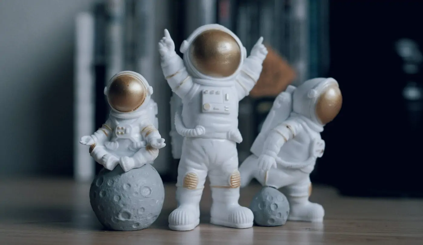 A photograph of three cartoonish astronauts figurines on a desk. One sits on a tiny moon in the lotus yoga pose, the next stands triumphantly with hands extended in the air, the last croquettes over as if taking a step in low gravity.