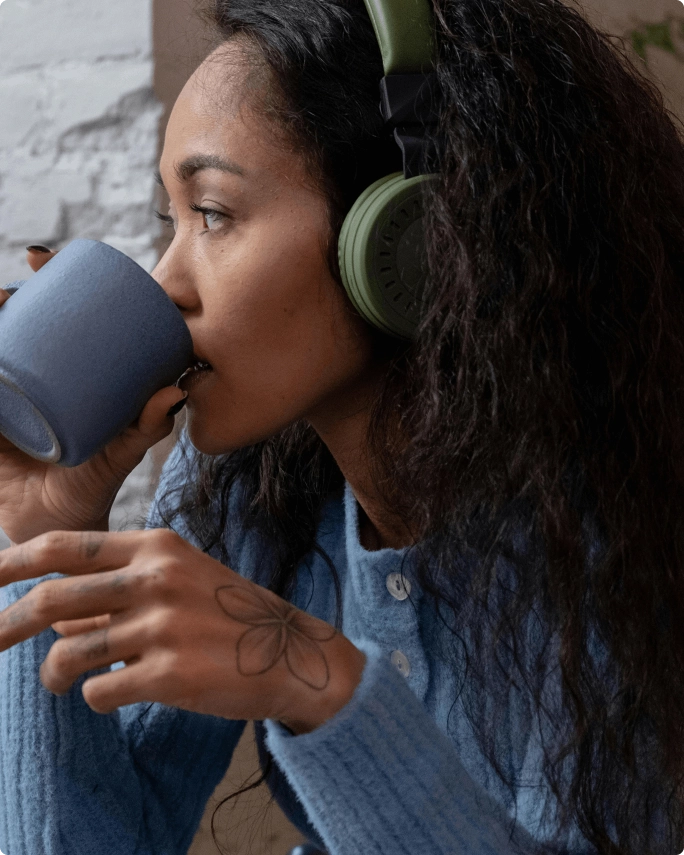 A woman wearing headphones, drinking from a mug.