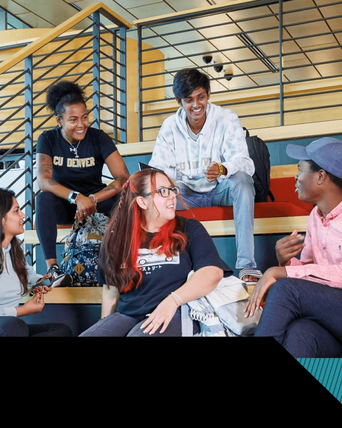 Diverse CU Denver students sit together on tiered seating inside a campus building, smiling and chatting in a relaxed, social atmosphere.
